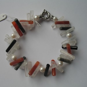 Könnte beinhalten: Ein handgefertigtes Armband mit einer Mischung aus transparenten, roten, schwarzen und grauen rechteckigen Perlen. Das Armband enthält auch weiße Perlen und einen silbernen Verschluss. Die Perlen sind in einem einzigartigen, auffälligen Muster angeordnet.