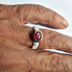 Könnte beinhalten: Ein silberner Ring mit einer strukturierten Band und einem roten ovalen Edelstein, der in einer goldenen Fassung sitzt.