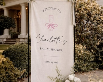 May include: A linen banner with the text "WELCOME TO Charlotte's BRIDAL SHOWER" and the date April 25, 2026. The banner is decorated with a champagne glass and bow graphic. The banner is displayed outdoors with floral arrangements.