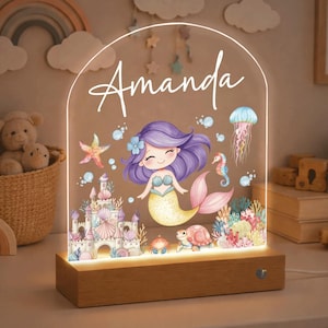 以下が含まれることがあります： 人魚のデザインと「Amanda」の名前が入ったパーソナライズされたナイトライト。アクリルパネルには、漫画の人魚、城、海の生き物が描かれており、木製のベースにセットされています。ライトは柔らかな光を放ちます。