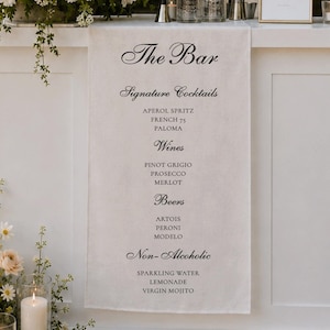 Puede incluir: Una barra blanca con arreglos florales y una pancarta que dice "The Bar". La barra ofrece cócteles de autor, vinos, cervezas y bebidas sin alcohol. Decorada con velas y cristalería.