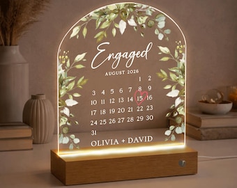 May include: A lighted acrylic engagement calendar with a wooden base. The calendar displays "Engaged" and "August 2026" with a highlighted date. Greenery and floral accents frame the top. The names "Olivia + David" are at the bottom.