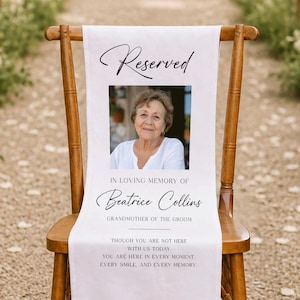 Può includere: Un cartello commemorativo in tessuto bianco drappeggiato su una sedia di legno. Presenta la parola Reserved, una fotografia di una donna, il testo In Loving Memory of Beatrice Collins, Grandmother of the Groom e una poesia sul ricordo.