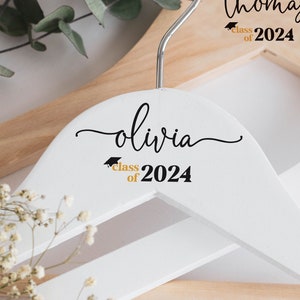 May include: Two white wooden hangers with black lettering. The top hanger says "Olivia class of 2024" and the bottom hanger says "Thomas class of 2024".