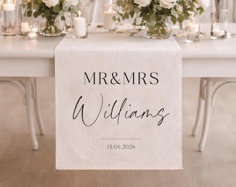 May include: A white table set for a wedding reception, with a floral centerpiece and candles. A white table runner displays the text "MR & MRS Williams" and the date 13.06.2026.