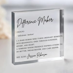 May include: A clear acrylic block with the words "Difference Maker" in elegant black script. The block features a definition of the term, including its pronunciation and two descriptive points. The name "Jessica Robinson" is also included.