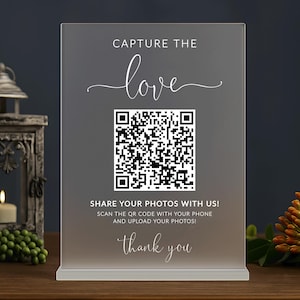 May include: A clear acrylic sign with the words "CAPTURE THE love" in elegant script. A QR code is centered below, with the text "SHARE YOUR PHOTOS WITH US!" and "thank you" at the bottom. The sign is on a wooden surface.