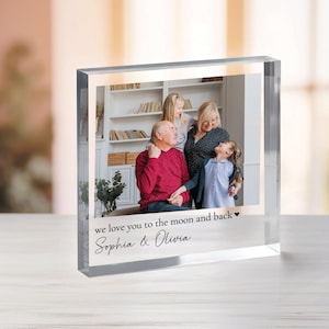 Può includere: Un cubo fotografico in vetro trasparente con una foto di famiglia all'interno. La foto mostra una famiglia di quattro persone, tra cui due bambine, un uomo e una donna. Il testo "we love you to the moon and back" è stampato sul fondo del cubo, insieme ai nomi "Sophia & Olivia".