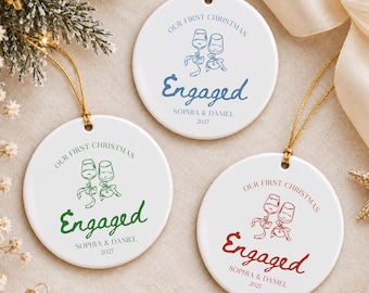 May include: Three round white ceramic ornaments with gold hanging strings. Each features a graphic of two wine glasses, the text "OUR FIRST CHRISTMAS", "Engaged", "SOPHIA & DANIEL", and "2027" in blue, green, or red ink respectively.