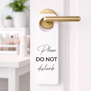 Może przedstawiać: Biały wieszak na drzwi z napisem "Please DO NOT disturb" czarnym skryptem. Wieszak wisi na drzwiach ze złotą klamką. Tło to rozmyty obraz białego stołu i roślin.