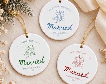 May include: Three round white ceramic ornaments with gold hanging strings. Each features a line drawing of two wine glasses, the text "OUR FIRST CHRISTMAS", "Married", "SOPHIA & DANIEL", and "2027" in blue, green, or red ink respectively.
