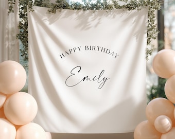 May include: A white fabric banner with the text "HAPPY BIRTHDAY Emily" in black script. The banner is decorated with greenery and surrounded by peach-colored balloons, creating a festive atmosphere for a birthday celebration.