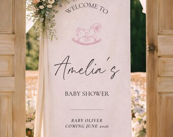 May include: A cream-colored baby shower sign with the text "Welcome to Amelia's Baby Shower." A pink rocking horse graphic is above the name. The sign also includes the text "Baby Oliver Coming June 2026."