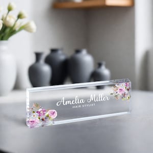 May include: Clear acrylic desk nameplate with a floral design and the text "Amelia Miller Hair Stylist".
