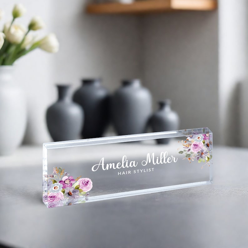 May include: Clear acrylic desk nameplate with a floral design and the text "Amelia Miller Hair Stylist".