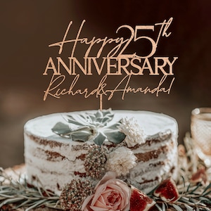 May include: A wooden cake topper with gold glitter, reading "Happy 25th Anniversary Richard & Amanda". The topper is on a white cake decorated with greenery and flowers.