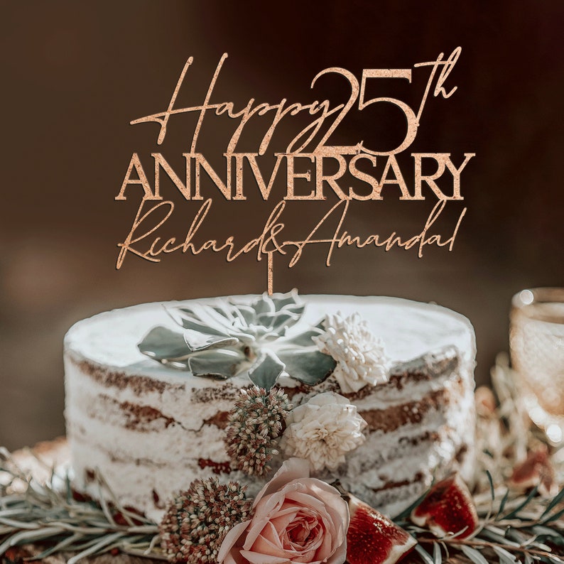 May include: A wooden cake topper with gold glitter, reading "Happy 25th Anniversary Richard & Amanda". The topper is on a white cake decorated with greenery and flowers.
