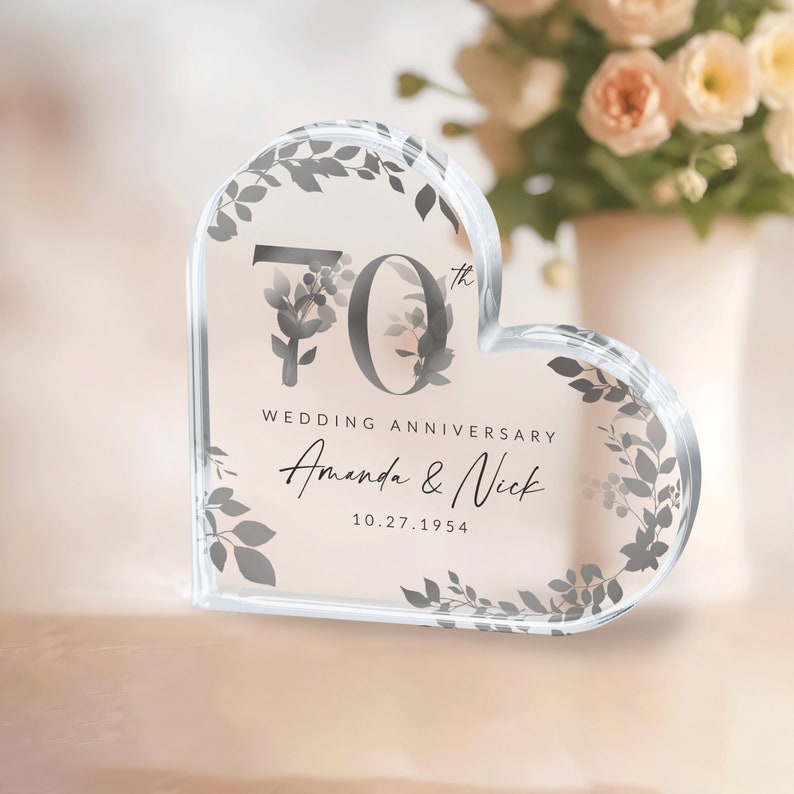 May include: A clear heart-shaped acrylic paperweight with a floral design and the text "70th Wedding Anniversary Amanda & Nick 10.27.1954"
