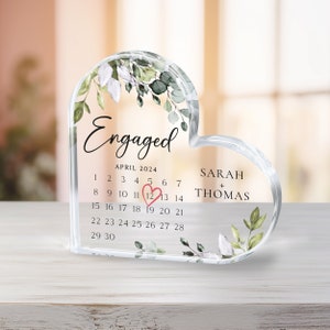 K&ouml;nnte beinhalten: Ein transparentes, herzf&ouml;rmiges Acryl-Andenken mit einem Blumendesign und dem Text "Verlobt" und "APRIL 2024". Das Datum "12" ist mit einem rosa Herzen hervorgehoben. Die Namen "SARAH + THOMAS" sind unter dem Kalender gedruckt.