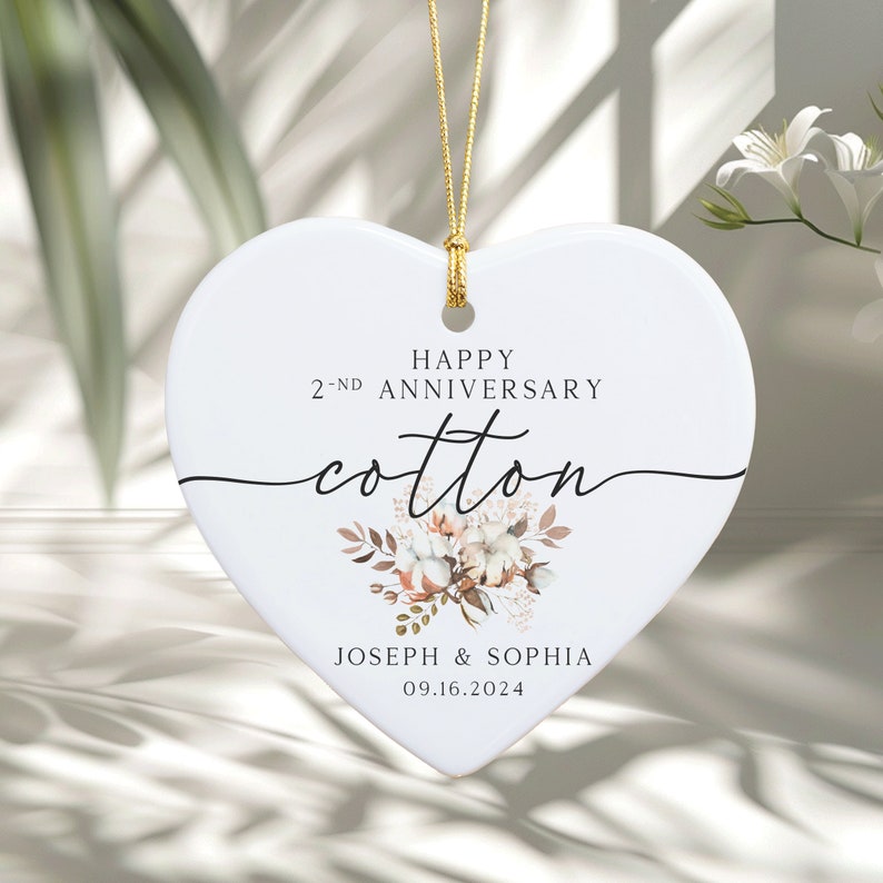 Peut inclure: Ornement en c&eacute;ramique blanche en forme de c&oelig;ur avec une ficelle dor&eacute;e pour l'accrocher. L'ornement pr&eacute;sente un motif floral avec des boules de coton et le texte "Happy 2nd Anniversary Cotton Joseph & Sophia 09.16.2024".
