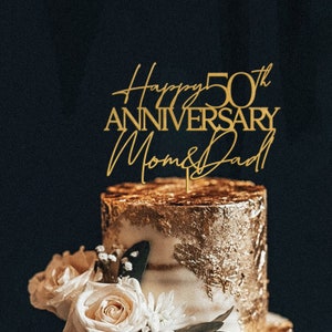 May include: Gold cake topper with the text "Happy 50th Anniversary Mom & Dad" on a two-tiered cake decorated with gold leaf and white frosting.