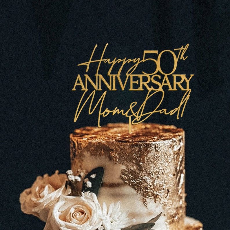 May include: Gold cake topper with the text "Happy 50th Anniversary Mom & Dad" on a two-tiered cake decorated with gold leaf and white frosting.