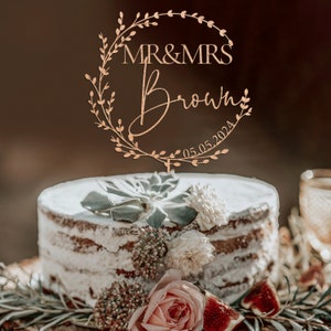K&ouml;nnte beinhalten: Ein h&ouml;lzerner Tortendekor mit einem Kranzdesign und dem Text "MR & MRS Brown 05.05.2024". Der Dekor befindet sich auf einem wei&szlig;en Kuchen, der mit Gr&uuml;npflanzen und Blumen verziert ist.