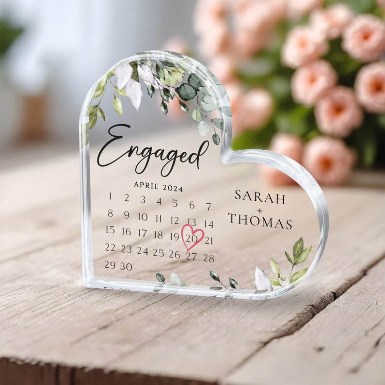 K&ouml;nnte beinhalten: Ein transparentes, herzf&ouml;rmiges Acryl-Andenken mit einem Blumendesign und dem Wort "Verlobt" oben. Der untere Teil des Herzens zeigt einen Kalender mit dem Datum "20", das in Rosa umkreist ist, und die Namen "Sarah + Thomas" darunter.