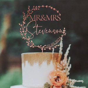 K&ouml;nnte beinhalten: Ein ros&eacute;goldener Hochzeitstortenaufsatz mit einem Design aus einem Bl&auml;tterkranz. Der Aufsatz tr&auml;gt den Text "MR & MRS Stevenson 05.05.2024".