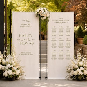 Op de afbeelding: Twee verticale off-white stoffen banners opgehangen aan zwarte metalen standaards. De linker banner toont WELCOME TO THE WEDDING OF HAILEY and THOMAS 13TH MARCH 2026 BELMONTE ESTATE, ITALY. De rechter banner bevat een lijst met tafelindelingen.