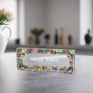 May include: A clear glass desk nameplate with a floral border and the text "Abigail Harris, Project Manager".
