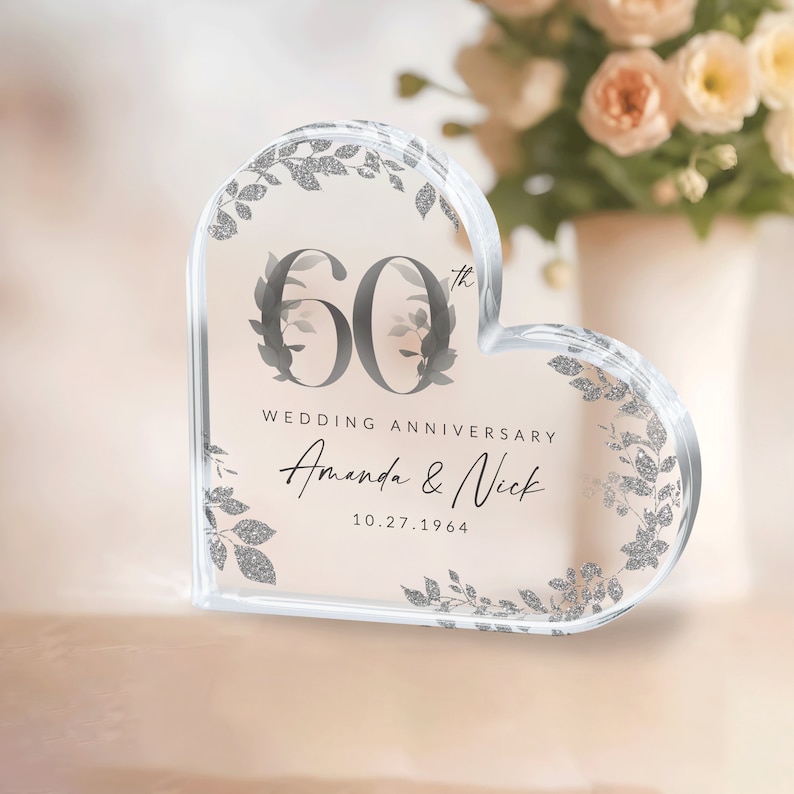May include: A clear heart-shaped crystal paperweight with a silver glitter design. The number 60 is in the center with the text "Wedding Anniversary" and the names "Amanda & Nick" and the date "10.27.1964".