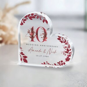 May include: A clear heart-shaped acrylic paperweight with a red glitter design. The paperweight features the number 40 with a small 'th' superscript, the text 'Wedding Anniversary', and the names 'Amanda & Nick' with the date '10.27.1984'.