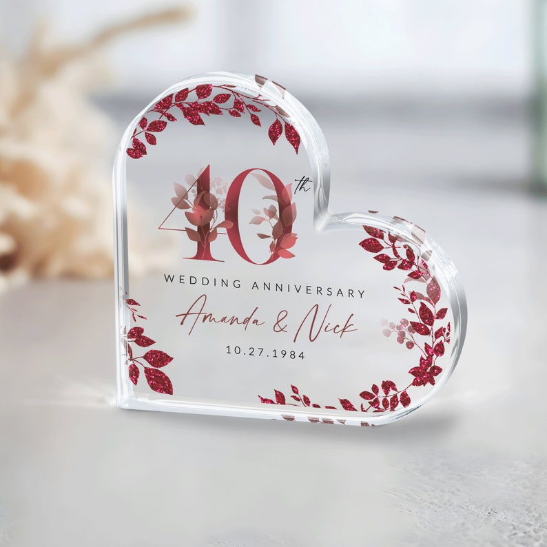May include: A clear heart-shaped acrylic paperweight with a red glitter design. The paperweight features the number 40 with a small 'th' superscript, the text 'Wedding Anniversary', and the names 'Amanda & Nick' with the date '10.27.1984'.
