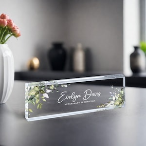 May include: A clear acrylic desk nameplate with a green and white floral design. The nameplate reads "Evelyn Davis, Veterinary Technician".