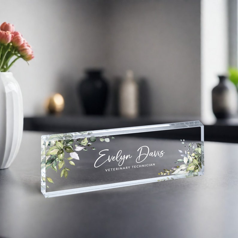 May include: A clear acrylic desk nameplate with a green and white floral design. The nameplate reads "Evelyn Davis, Veterinary Technician".