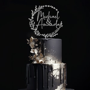 K&ouml;nnte beinhalten: Ein wei&szlig;er Holz-Kuchentopper mit einem Kranz-Design und dem Text "Michael & Amanda 05.05.2024". Der Topper befindet sich auf einer schwarzen und goldenen Hochzeitstorte.