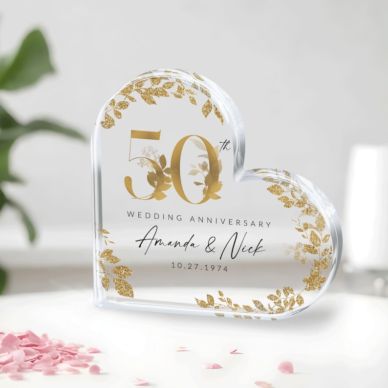 May include: A clear heart-shaped acrylic paperweight with gold glitter and floral accents. The number 50th is displayed in gold with the text "Wedding Anniversary Amanda & Nick 10.27.1974".