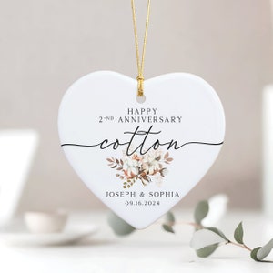 Peut inclure: Ornement en c&eacute;ramique blanc en forme de c&oelig;ur avec une ficelle dor&eacute;e pour l'accrocher. Il pr&eacute;sente un motif floral avec des boules de coton et le texte "Happy 2-nd Anniversary Cotton" et "Joseph & Sophia 09.16.2024".