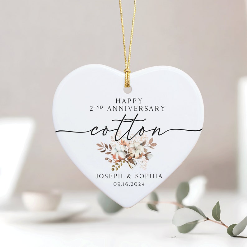 Peut inclure: Ornement en c&eacute;ramique blanc en forme de c&oelig;ur avec une ficelle dor&eacute;e pour l'accrocher. Il pr&eacute;sente un motif floral avec des boules de coton et le texte "Happy 2-nd Anniversary Cotton" et "Joseph & Sophia 09.16.2024".