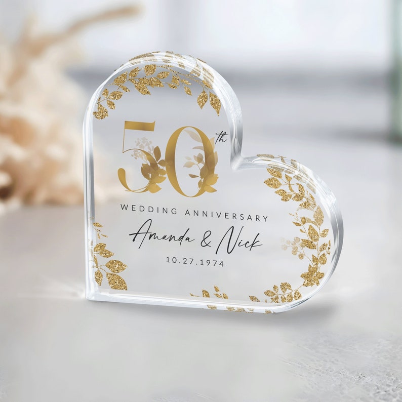 May include: A clear heart-shaped acrylic paperweight with gold glitter and floral accents. The number 50 with a 'th' superscript is in the center, along with the text 'Wedding Anniversary Amanda & Nick 10.27.1974'.
