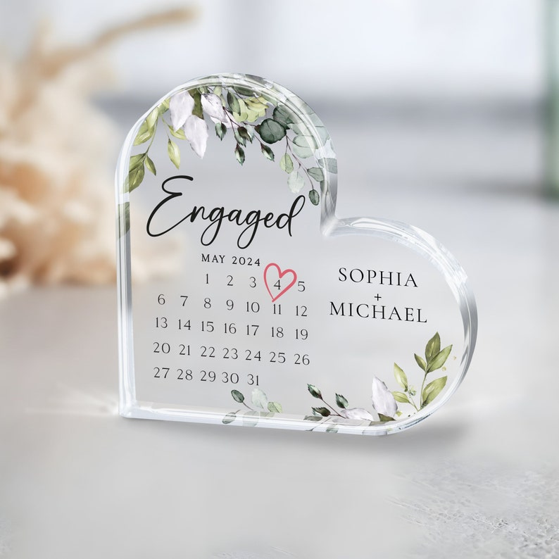 K&ouml;nnte beinhalten: Ein durchsichtiger, herzf&ouml;rmiger Kristall-Briefbeschwerer mit einem Blumenmuster und dem Text "Verlobt" oben. Der Briefbeschwerer enth&auml;lt auch einen Kalender f&uuml;r Mai 2024, wobei die Zahl 4 in Rosa hervorgehoben ist. Die Namen "Sophia + Michael" sind unter dem Kalender gedruckt.