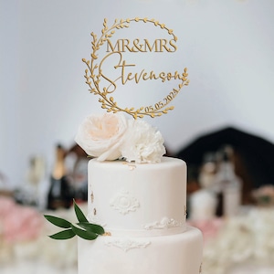 K&ouml;nnte beinhalten: Eine zweist&ouml;ckige wei&szlig;e Hochzeitstorte mit einem goldenen Kranz-Topper, auf dem "MR & MRS Stevenson 05.05.2024" steht. Die Torte ist mit wei&szlig;en Blumen und goldenen Akzenten verziert.