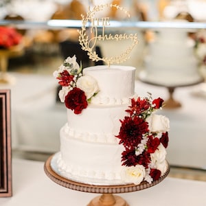 K&ouml;nnte beinhalten: Eine dreist&ouml;ckige wei&szlig;e Hochzeitstorte, verziert mit roten und wei&szlig;en Blumen. Die oberste Etage hat einen goldenen Topper mit der Aufschrift "The Johnsons" mit einem Kranz und dem Datum "12.12.2023".