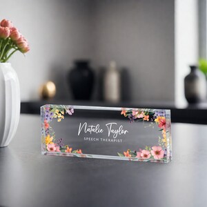 May include: A clear glass desk nameplate with a floral design in shades of pink, yellow, and purple. The name "Natalie Taylor" and the title "Speech Therapist" are printed in white on the nameplate.