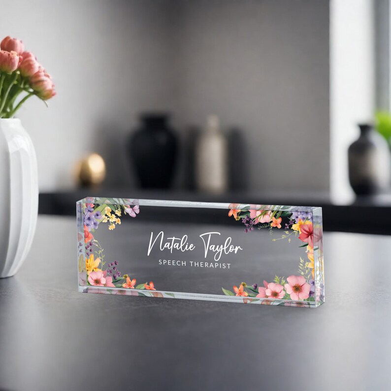 May include: A clear glass desk nameplate with a floral design in shades of pink, yellow, and purple. The name "Natalie Taylor" and the title "Speech Therapist" are printed in white on the nameplate.