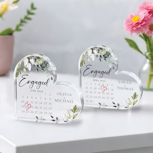 K&ouml;nnte beinhalten: Zwei transparente, herzf&ouml;rmige Acryl-Ornamente mit Blumendesigns und dem Wort "Verlobt" auf jedem. Die Ornamente zeigen Kalender f&uuml;r Juni und April 2024 mit den in Pink eingekreisten Daten. Die Namen Olivia + Michael und Sarah + Thomas sind auf den Ornamenten aufgedruckt.