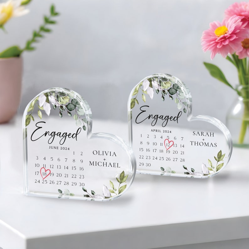 K&ouml;nnte beinhalten: Zwei transparente, herzf&ouml;rmige Acryl-Ornamente mit Blumendesigns und dem Wort "Verlobt" auf jedem. Die Ornamente zeigen Kalender f&uuml;r Juni und April 2024 mit den in Pink eingekreisten Daten. Die Namen Olivia + Michael und Sarah + Thomas sind auf den Ornamenten aufgedruckt.
