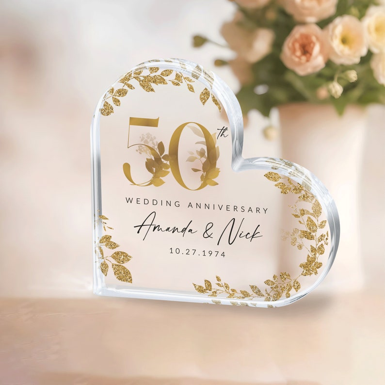 May include: A clear heart-shaped acrylic paperweight with gold glitter accents and a gold inscription that reads "50th Wedding Anniversary Amanda & Nick 10.27.1974".