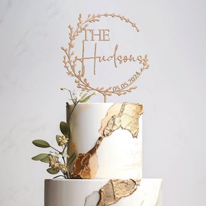 K&ouml;nnte beinhalten: Eine zweist&ouml;ckige Hochzeitstorte, verziert mit Blattgold und Gr&uuml;n. Die oberste Etage hat einen h&ouml;lzernen Tortendecker mit einem Kranzdesign und dem Text "THE Hudsons 05.05.2024".
